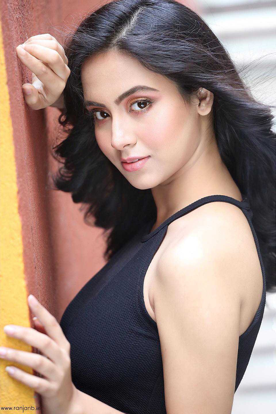 A striking portrait of Irin Chakraborty posing elegantly against a textured wall, wearing a black sleeveless top. Her confident gaze and flowing black hair create a captivating image. Photographed by Ranjan Bhattacharya Photography.