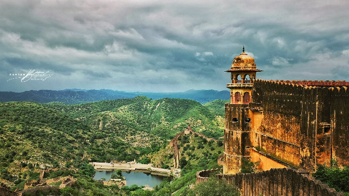 A panoramic view of an ancient fortress with a prominent tower overlooking a lush green valley and a body of water under a dramatic cloudy sky. Photo by Ranjan Bhattacharya.