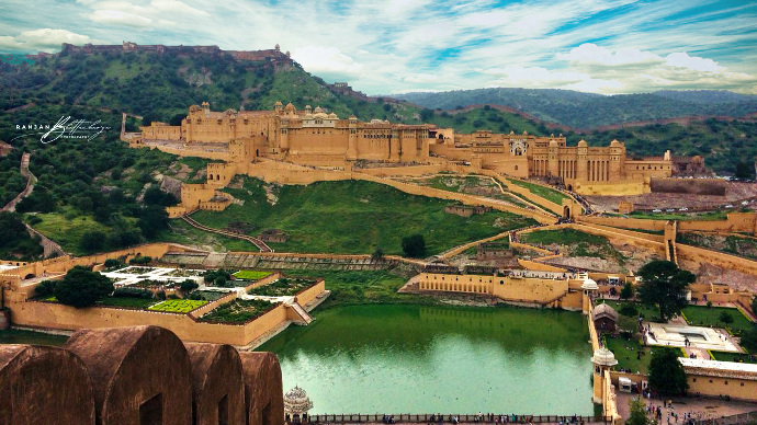 A panoramic view of a large, historic fort complex situated on a hill, surrounded by greenery and a large body of water in the foreground. Photo by Ranjan Bhattacharya.