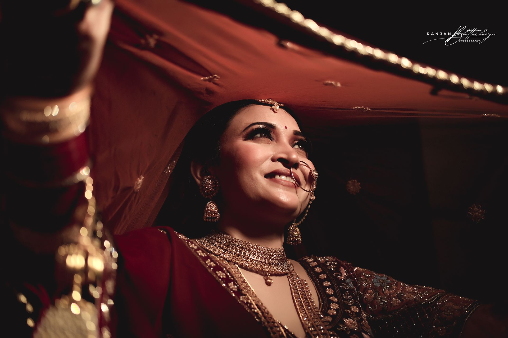 A joyful Ruhi Singh smiles under her bridal veil, adorned in intricate jewelry and a maroon bridal outfit, beautifully photographed by Ranjan Bhattacharya.