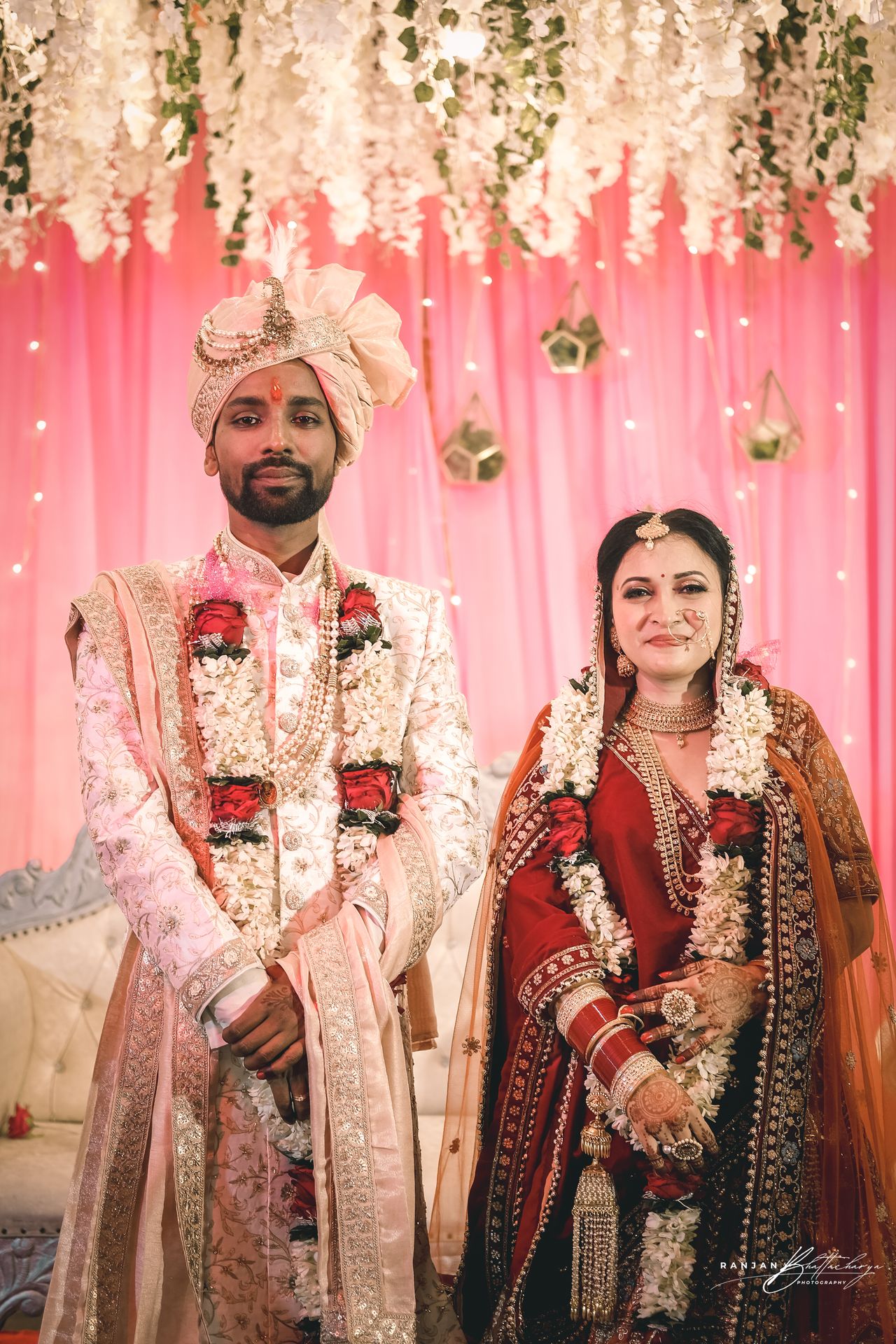 A wedding photograph featuring the bride, Ruhi Singh, and the groom, Roshan Kumar, in traditional Indian attire during a ceremony. The bride is adorned in an ornate orange saree with intricate embroidery, while the groom is dressed in a cream sherwani with pink detailing. They are standing under a floral canopy with white flowers and strings of lights, creating a festive and elegant atmosphere. The image captures a moment where the groom appears to be placing a garland around the bride’s neck, symbolizing their union. Photography by Ranjan Bhattacharya, captured in Bihar.