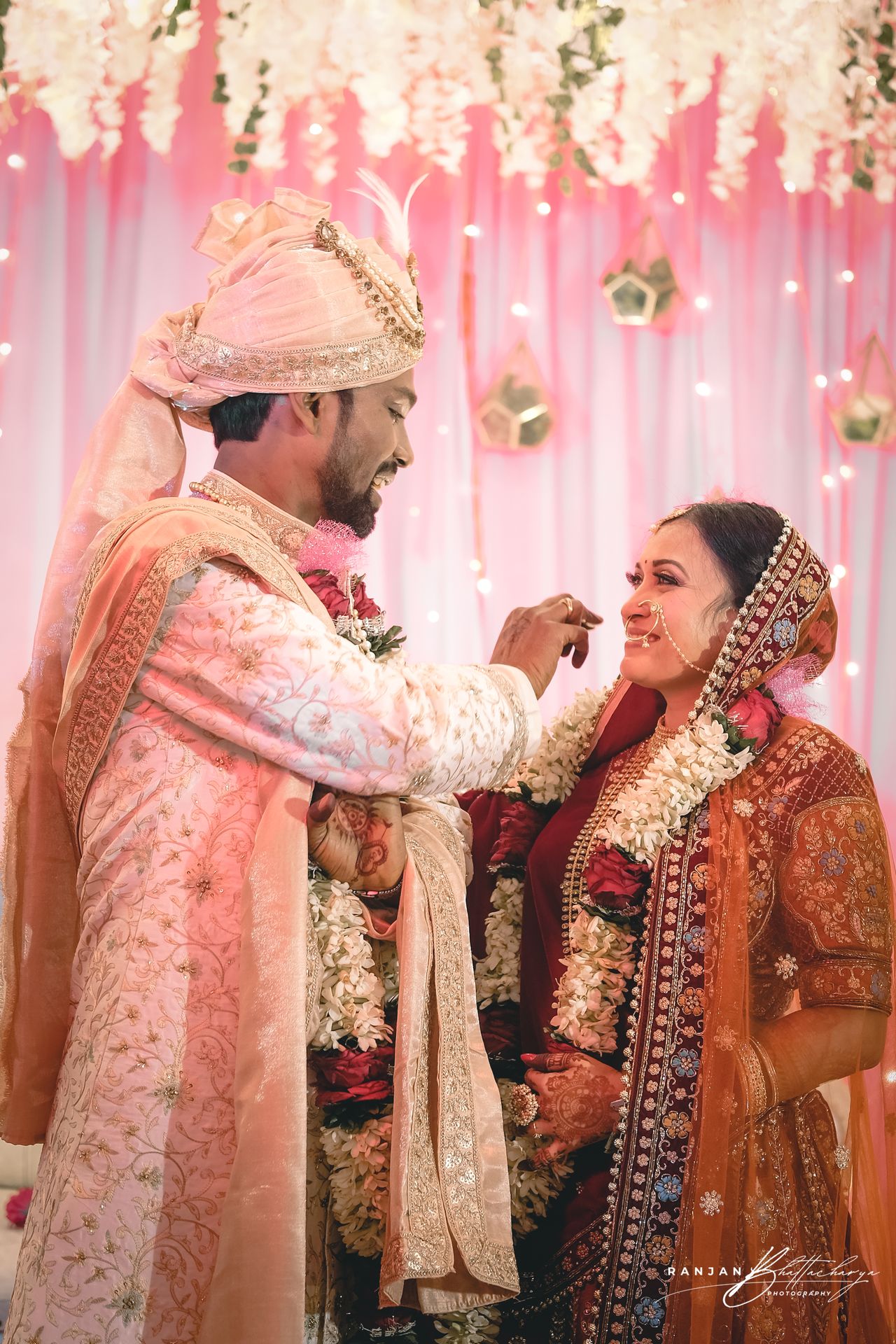 A wedding photograph featuring the bride, Ruhi Singh, and the groom, Roshan Kumar, in traditional Indian attire during a ceremony. The bride is adorned in an ornate orange saree with intricate embroidery, while the groom is dressed in a cream sherwani with pink detailing. They are standing under a floral canopy with white flowers and strings of lights, creating a festive and elegant atmosphere. The image captures a moment where the groom appears to be placing a garland around the bride’s neck, symbolizing their union. Photography by Ranjan Bhattacharya, captured in Bihar.