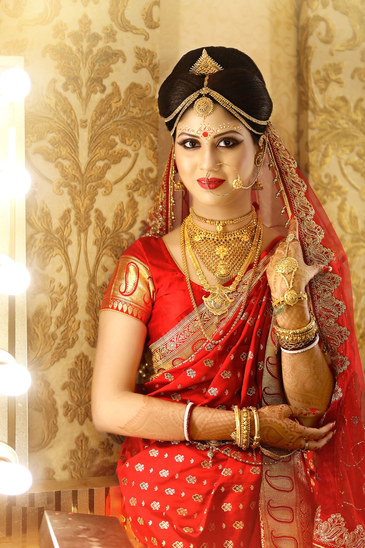 Bride Debarati Sarkar in traditional bridal attire and jewelry, getting final touches on her hairstyle by hands adorned with red bangles, against a softly blurred warm-lit background.