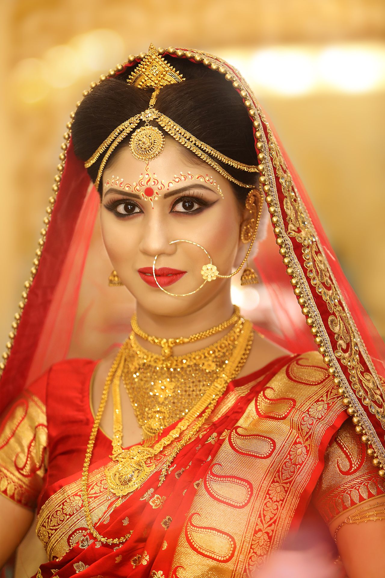 Bride Debarati Sarkar in traditional bridal attire and jewelry, getting final touches on her hairstyle by hands adorned with red bangles, against a softly blurred warm-lit background.