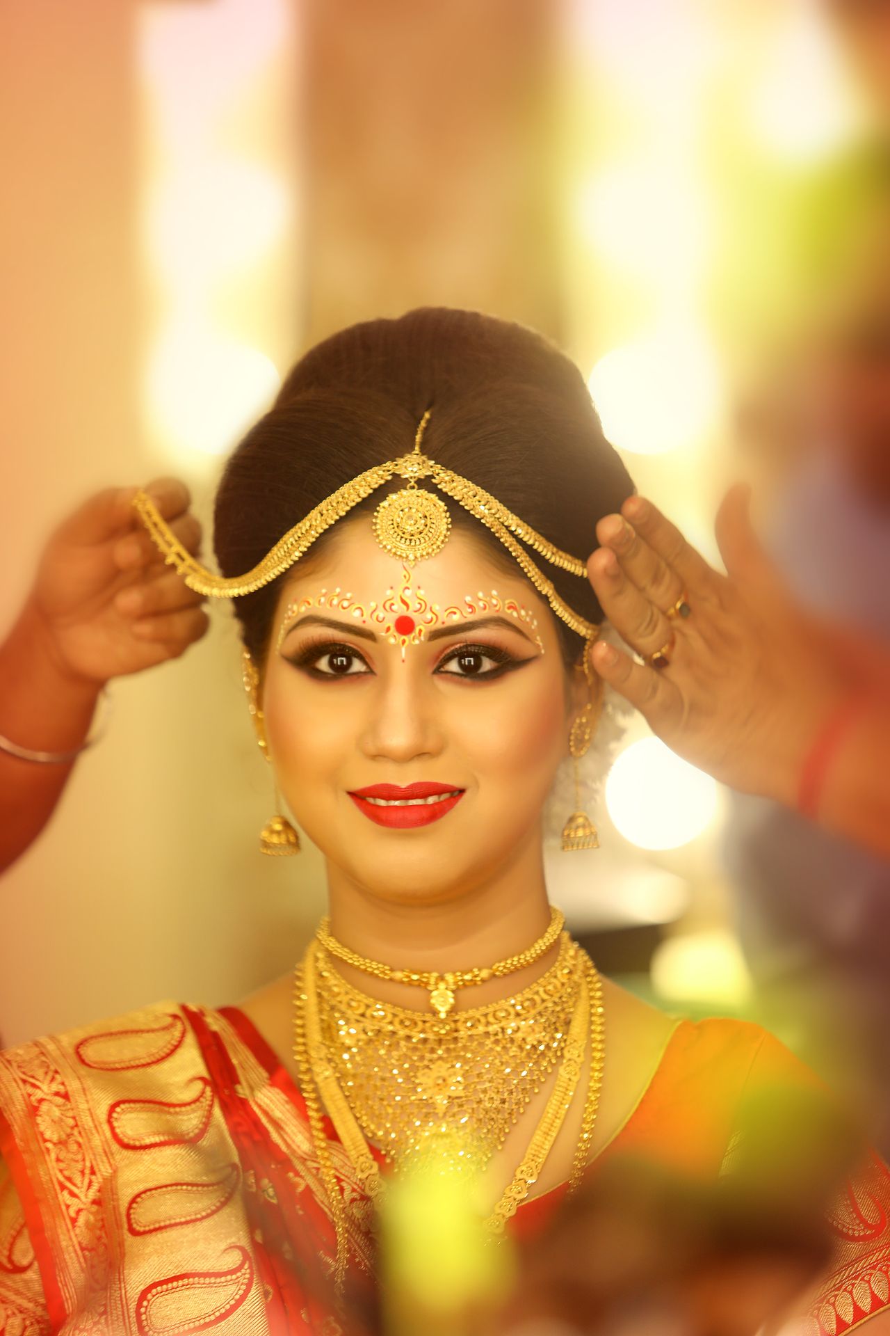 Bride Debarati Sarkar in traditional bridal attire and jewelry, getting final touches on her hairstyle by hands adorned with red bangles, against a softly blurred warm-lit background.