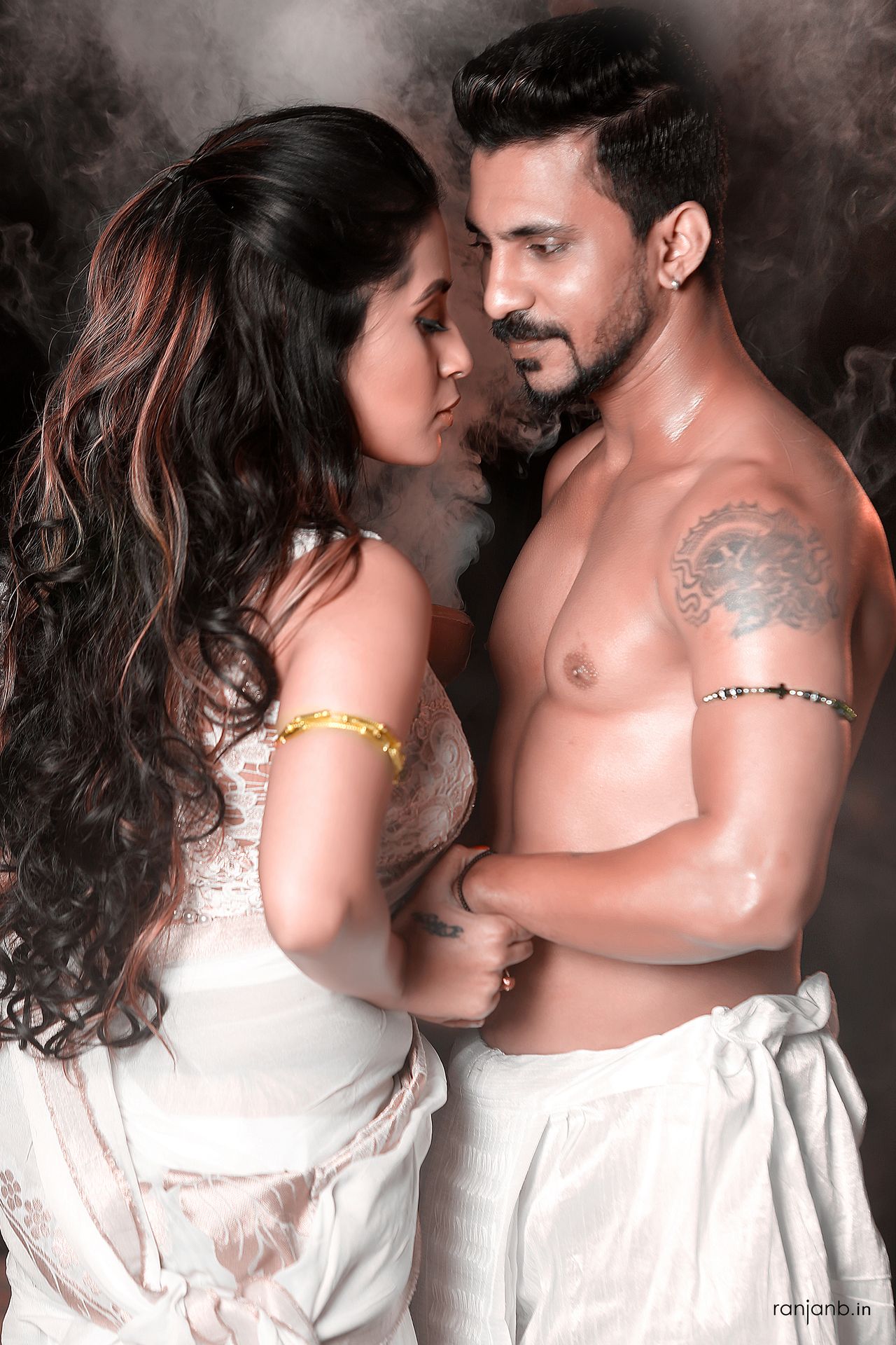 A couple intimately posing against a smoky backdrop, with the woman in a white saree and the man shirtless, showcasing their deep connection during their pre-wedding shoot.