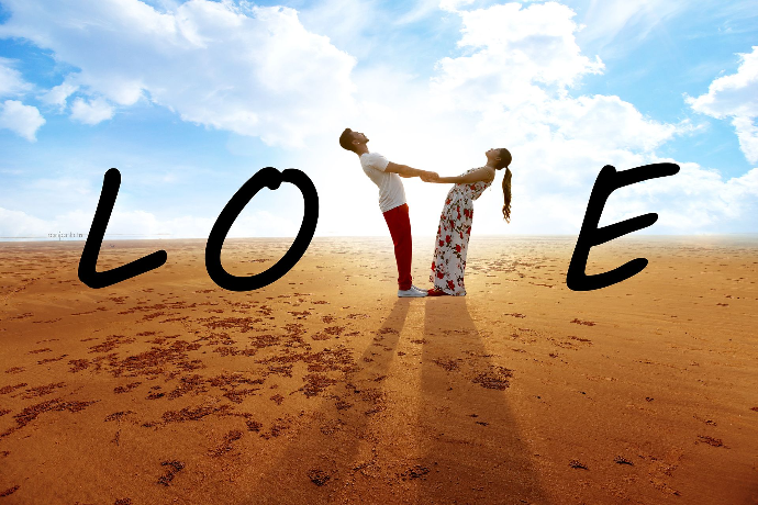 A couple standing on a vast, sunlit beach with the word 'LOVE' written around them, holding hands and leaning backward during their pre-wedding shoot.