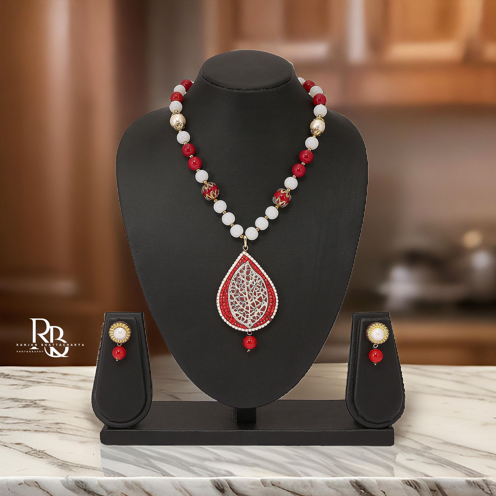 A red and white beaded necklace set with a large red pendant and matching earrings displayed on a black jewelry stand, photographed by Ranjan Bhattacharya.