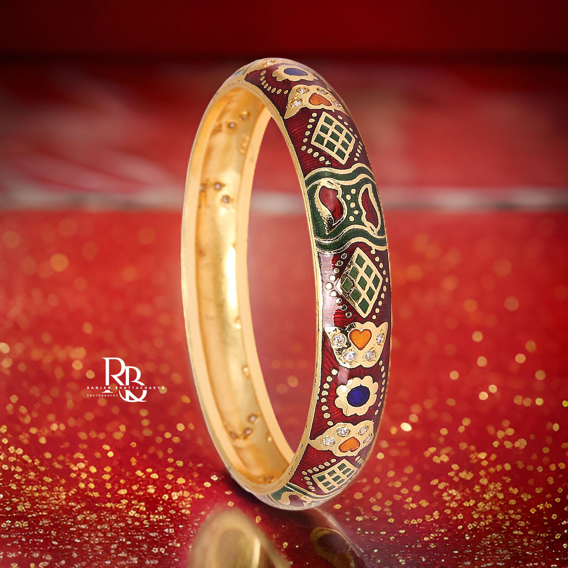 A traditional gold bangle with detailed enamel work in red, green, and gold colors, embellished with intricate patterns and hearts, photographed by Ranjan Bhattacharya.