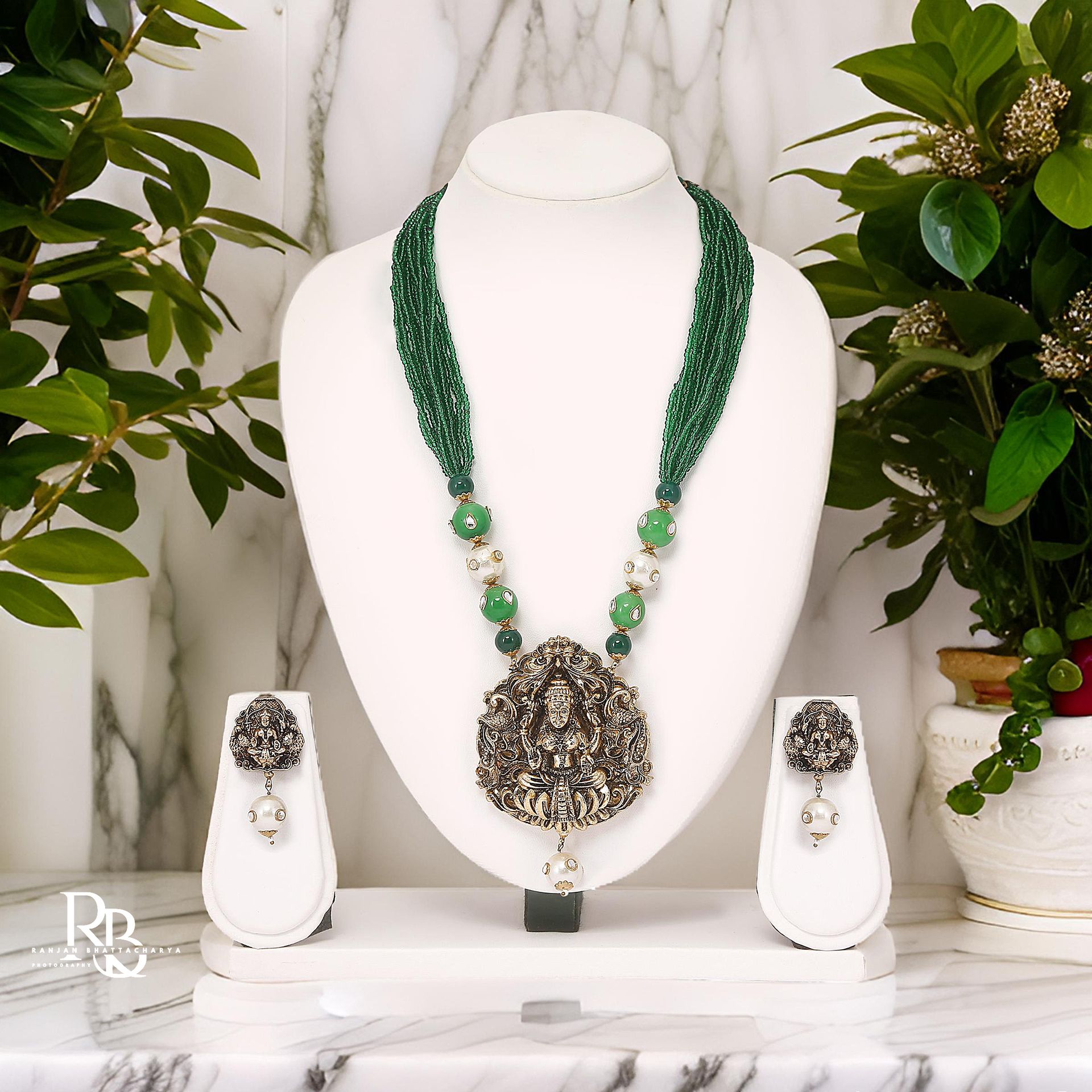 A green beaded necklace with a silver Ganesha pendant and matching earrings displayed on a white jewelry stand, photographed by Ranjan Bhattacharya.