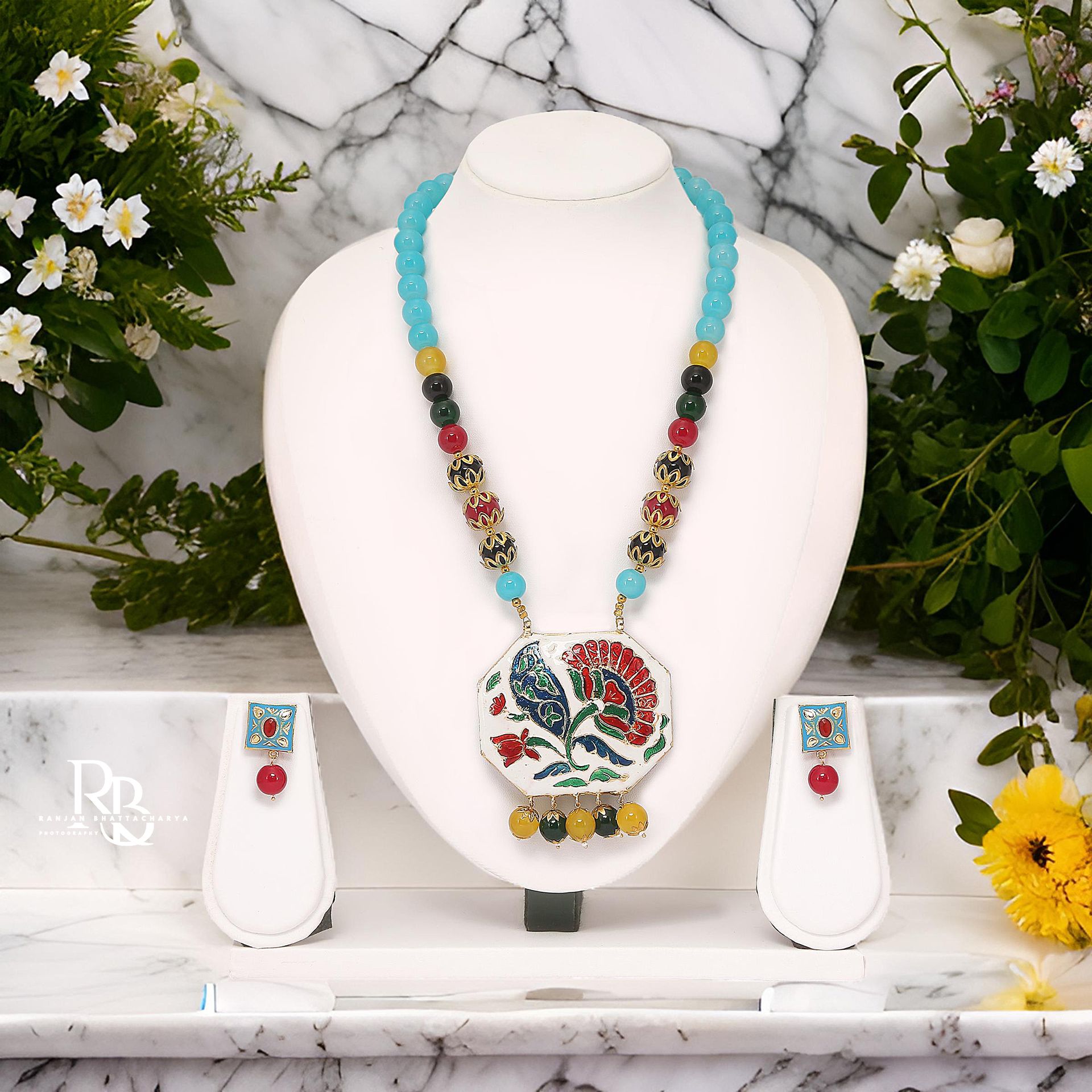 A beautiful necklace with blue and colorful beads and an artistic pendant with floral design displayed on a white mannequin, photographed by Ranjan Bhattacharya Photography.