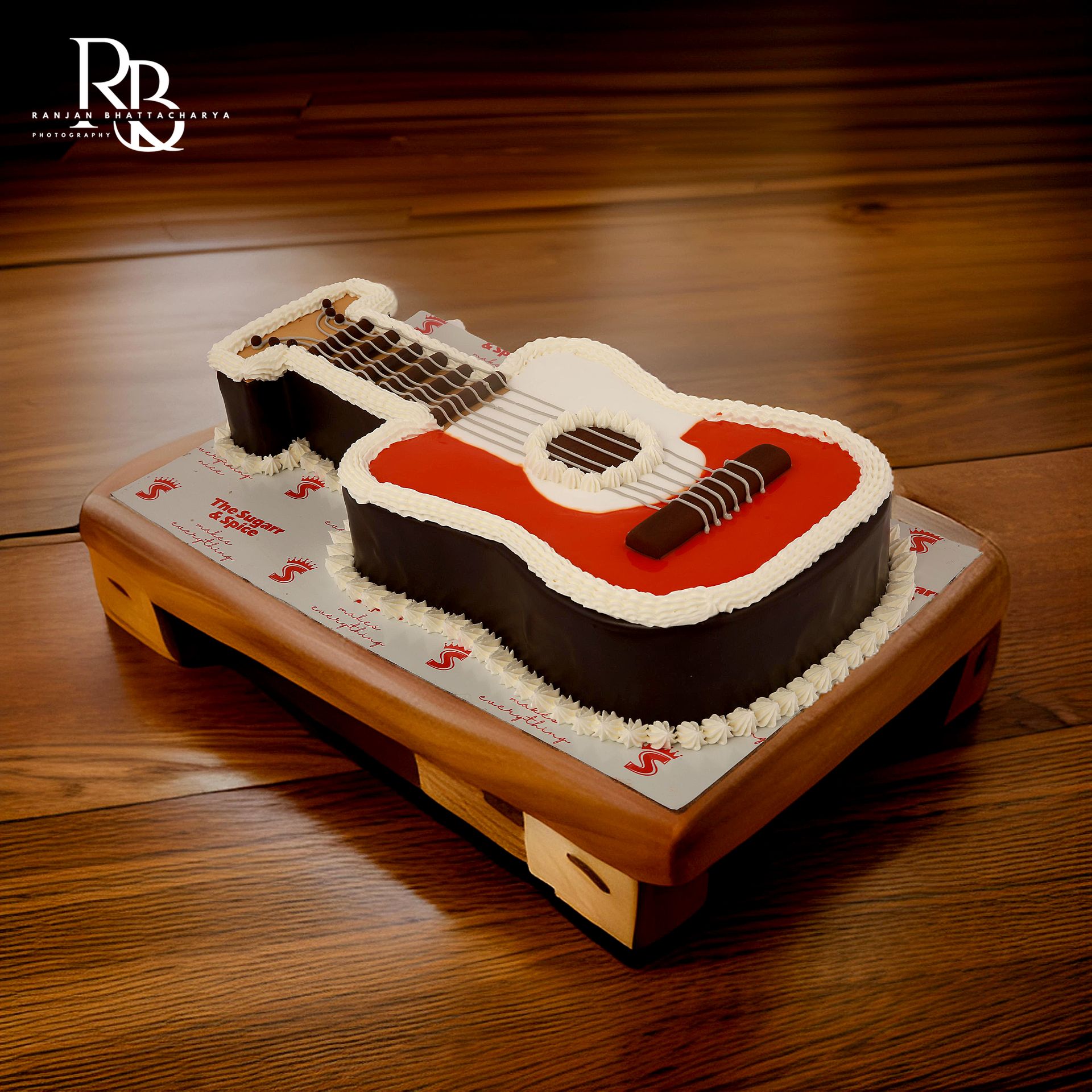 A guitar-shaped cake made of chocolate and white cream, captured on a wooden table by Ranjan Bhattacharya Photography.