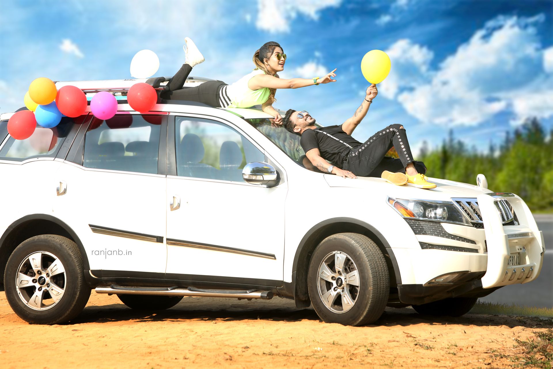 A couple posing playfully on top of a white SUV, decorated with colorful balloons, during their pre-wedding shoot. The woman is lying on the roof with her hand reaching out, while the man lies on the hood holding a yellow balloon.