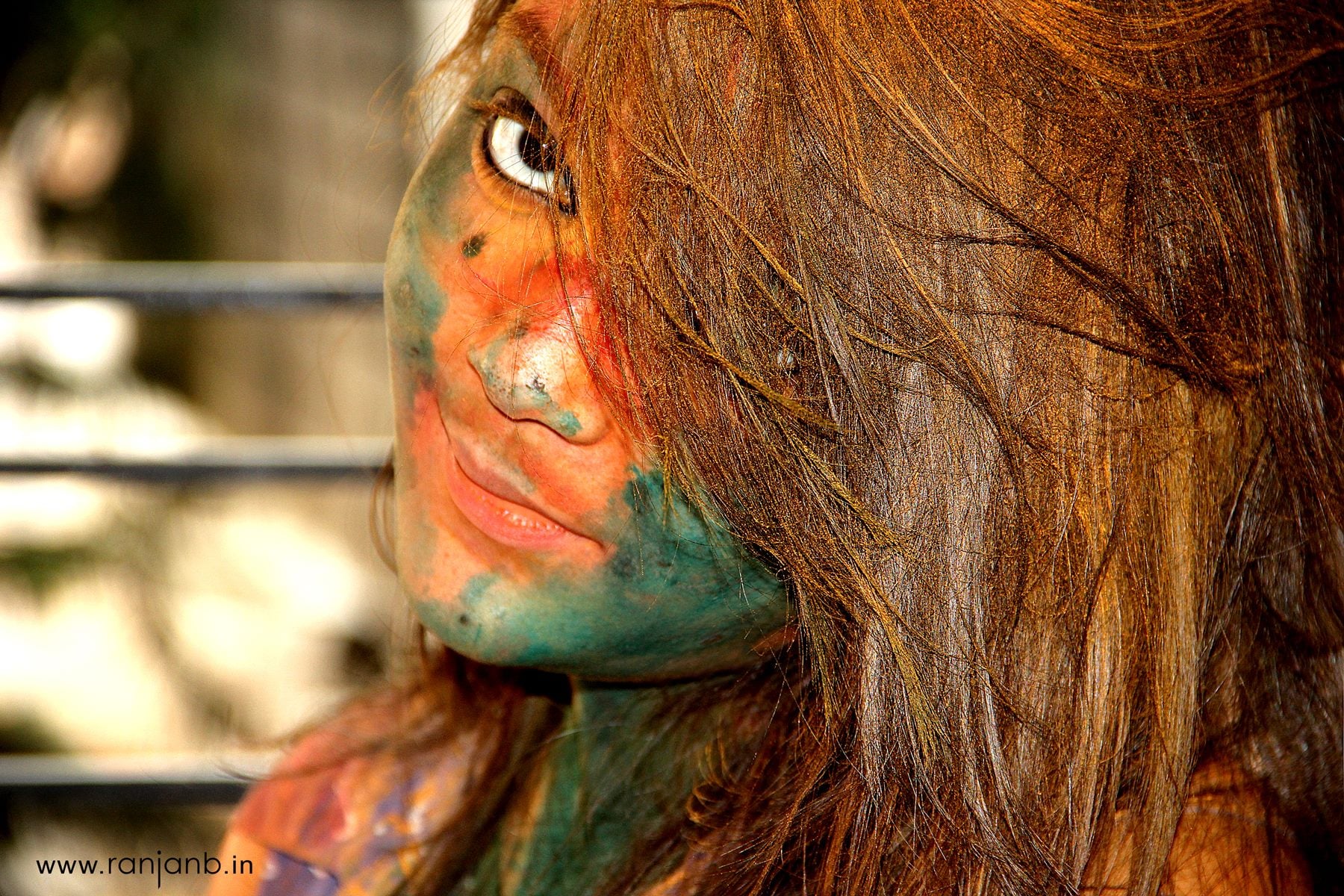 Close-up portrait of Camellia Roy during the vibrant Holi festival, captured by Ranjan Bhattacharya Photography. Her face is adorned with splashes of green and orange colors, and sunlight highlights the textures and hues on her skin and hair.